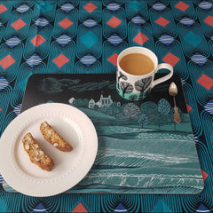 A snack of  2 biscotti and a mug of coffee on a mat with a landscape print in dark blue-greens on an african print table cloth
