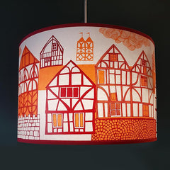 Lush Designs lampshade printed with a picture of old-fashioned buildings in red and orange, pictured against a very dark blue wall