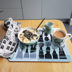 fruit topped meusli in a bowl with mug with goose print, small green jug and house-print napkin on a mat with tree design print