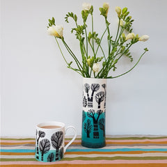 vase printed with black winter trees with a dipped turquoise glaze , filled with white freesias next to a mug with the same tree print 