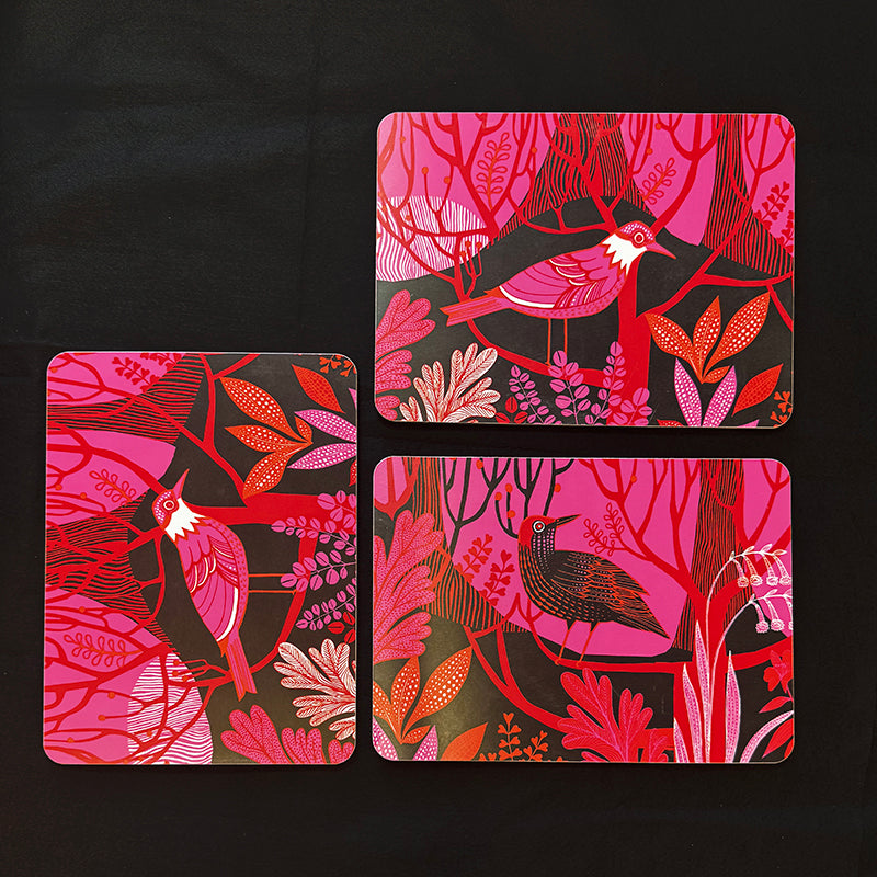 Three rectangular table mats with rounded corners arranged on a black background, featuring a vibrant pink, red, and orange botanical and bird illustration.