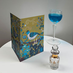 Decorative card with a bird design next to a glass of blue liquid and a perfume bottle on a white surface.
