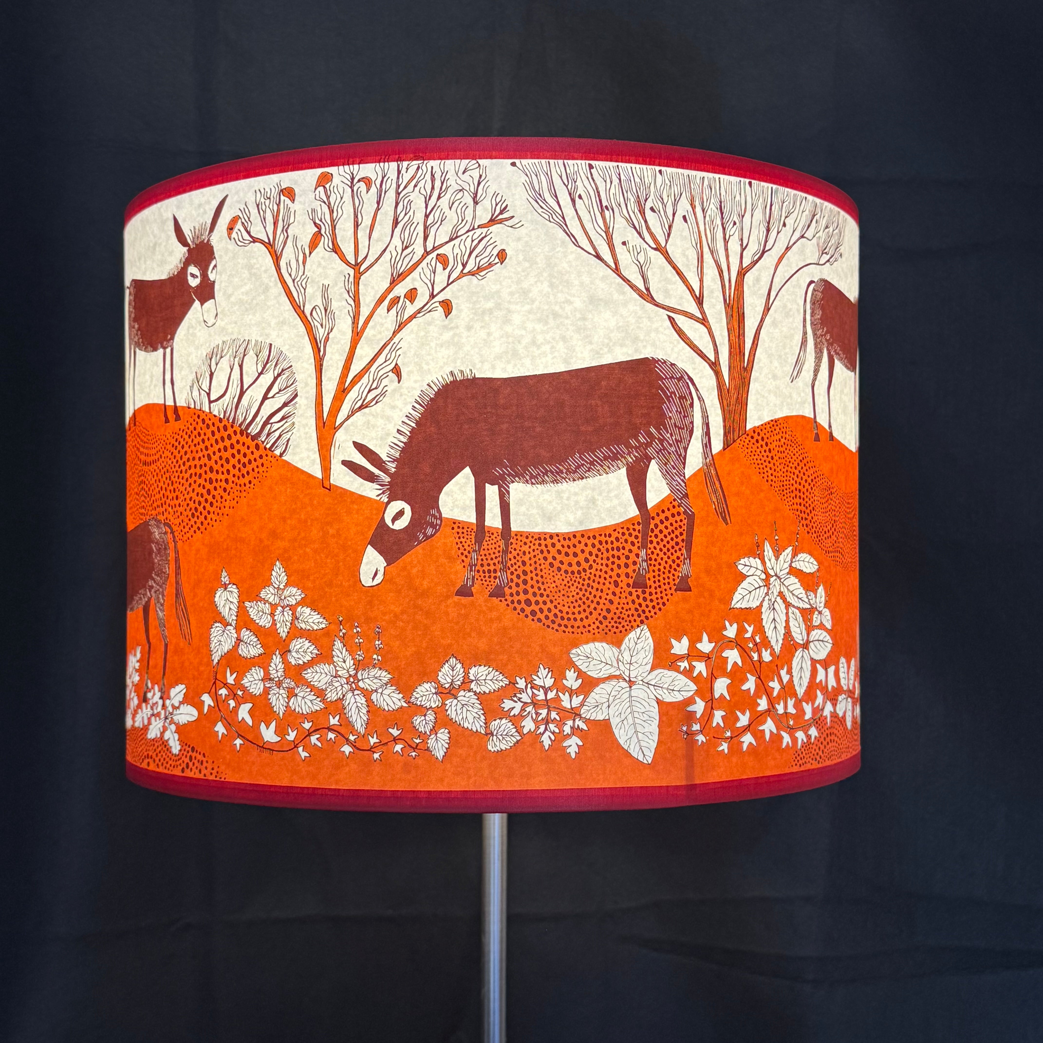 A cylindrical table lamp shade with a printed woodland scene featuring donkeys, trees, and white flowers on an orange and cream background, with red trim, shown against a dark backdrop.