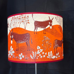 A cylindrical table lamp shade featuring a whimsical donkey illustration pattern in orange, cream, and dark brown, set against a black background.