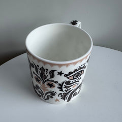 White mug with black and brown paisley pattern on a white surface with gray background