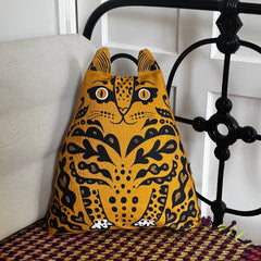 A mustard yellow cat-shaped fabric bag with black patterned details and white eyes, resting on a woven surface next to a light-colored cushion in front of a white paneled wall and black metal bed frame.