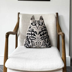 A mid-century modern wooden armchair with off-white textured fabric upholstery, featuring a black and white cat-patterned throw pillow on the seat, set in a room with gray herringbone tile flooring.