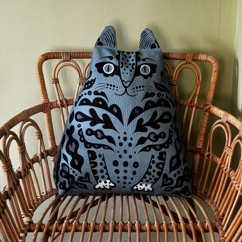 A blue cat-shaped decorative pillow with black patterns and white eyes, resting on a brown wicker chair against a light green wall.