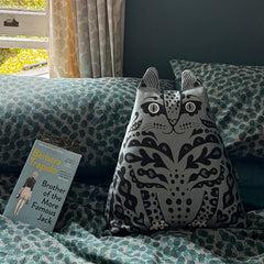 A blue and black cat-shaped throw pillow with embroidered details sits on a teal and white patterned bedspread next to a book.
