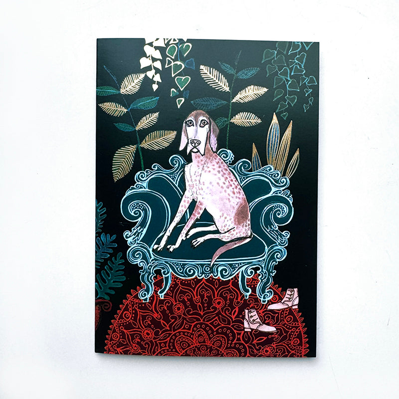 A card featuring a whimsical illustration of a spotted pink dog sitting on an ornate blue chair against a dark background, with decorative leaves, red patterned ground, and small shoes at the bottom, photographed on a white surface.