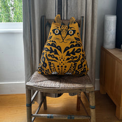 A yellow and black patterned cat-shaped throw pillow sits on a rustic wooden chair in a home interior setting.