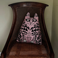 A pink throw pillow with a black cat pattern, resting on the back of a dark brown wooden cane chair.
