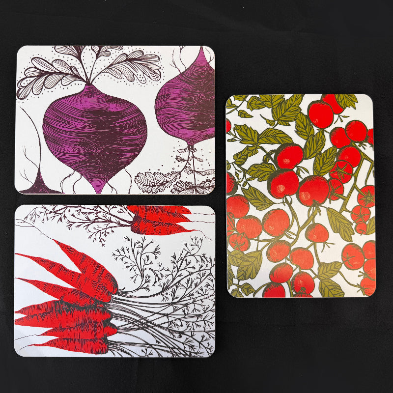 Three rectangular printed mats with botanical illustrations arranged on a black background. One shows purple beets with gray roots, one shows red carrots with gray tops, and one shows red tomatoes with green leaves on a white base.