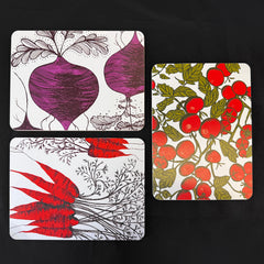 Three rectangular printed mats with botanical illustrations arranged on a black background. One shows purple beets with gray roots, one shows red carrots with gray tops, and one shows red tomatoes with green leaves on a white base.