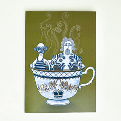 A card featuring a whimsical illustration of two stylized figures inside an ornate teacup on an olive green background, photographed against a plain off-white wall.