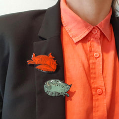 close up of enamel fox badge and cat pin on black lapel blazer worn with orange-red shirt