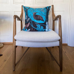 Shades of blue, black and brown cushion printed with a thrush bird in woodland setting on an easy chair made of wood with linen upholstery