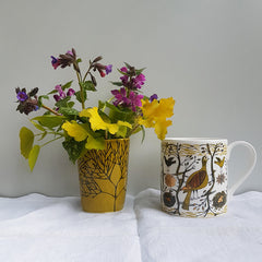 Mustard coloured ceramic beaker with posy of purple spring flowers next to chine mug printed with mustard, black and gold lustre birds and trees