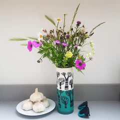 Lush Designs cylindrial vase printed with winter trees and dipped turquoise glaze, containing a selction of purple and yellow wild flowers next to a plate with garlic on it