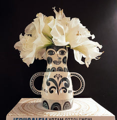 Lady-shaped vase with handles like arms with large white flowers in it that look like an extravagant hat