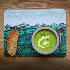 Mat depicting landscape of green hills with green pea soup and toast and a pewter spoonst