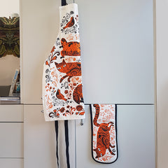 apron and bag hanging on a door, both printed with pattern of orange and black patterned cats, flowers and birds