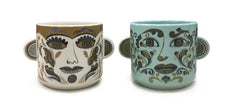 Two ceramic plant pots that look like heads printed with faces with ears sticking out the sides, one cream with black and olive print, one glazed in jade green