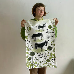 Smiling woman holding up a tea towel printed with donkeys in a flowery meadow