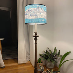 Lush Designs lampshade with print of a coastal village and the sea and sky in shades of blue shown in a room on a tall wooden lamp