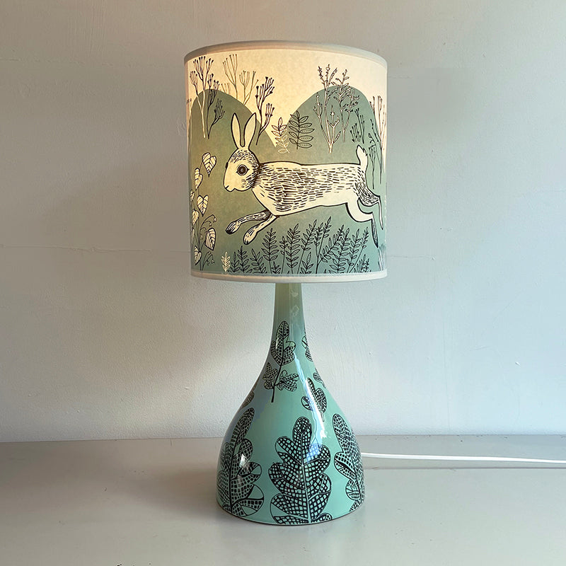 Lush Designs pale turquoise-green lamp base with black print of leaves paired with soft blue-green shade with print of a running rabbit 
