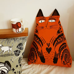 Lush Designs cat-shaped cushion in rich orange pictured on a sofa next to a lion-print cup