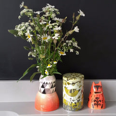 Orange kitty jar pictured with siskin bird-printed storage tin and orange and black patterned vase of daisies